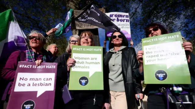 Women hold flags and signs saying "women are adult human females" in an outdoor demonstration.