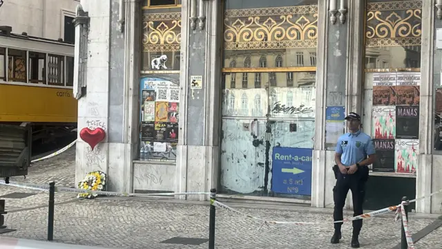 A police officer stands guard outside the cordon at the base of the funicular, where one carriage sits. A wreath of flowers with a red heart-shaped balloon sits against a nearby wall
