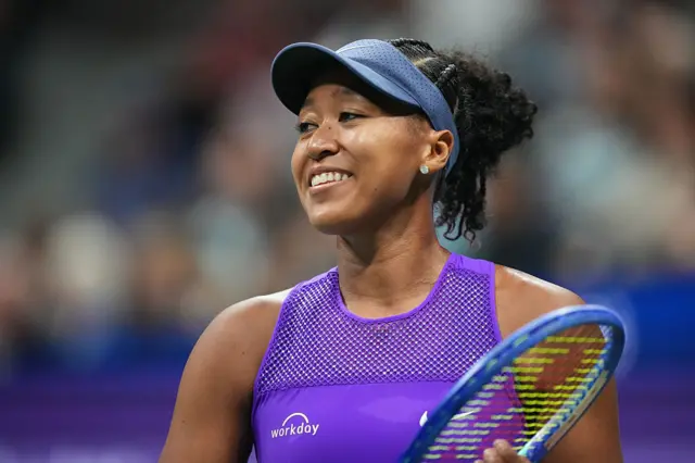 Naomi Osaka smiles and holds a tennis racquet