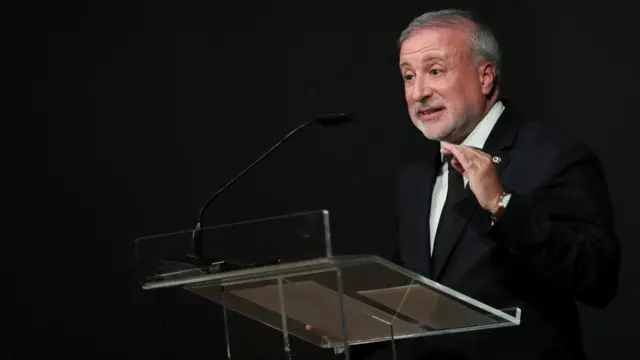 Pedro de Brito Bogas, wearing a black suit and tie, stands against a black backdrop speaking at a glass podium