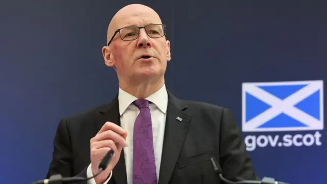 John Swinney, a bald man with glasses, speaks at a podium in front of a blue Scottish government sign. He is wearing a dark suit, white shirt and purple tie.