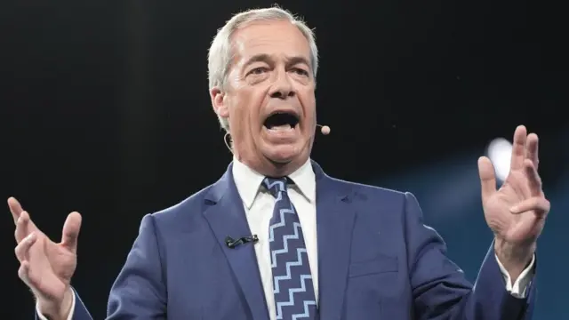 Farage speaks on stage, wearing a suit.