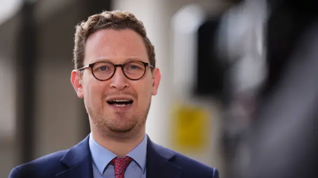A headshot of Darren Jones wearing a suit mid-speaking. The background is blurred
