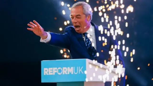 Farage wears a suit and gestures with his hand standing behind a podium.