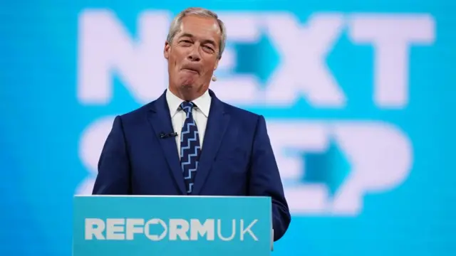 Farage smirks standing behind a podium wearing a suit.