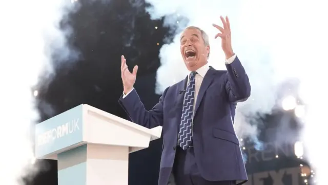 Farage smiles and gestures with his hands behind a podium reading Reform UK.