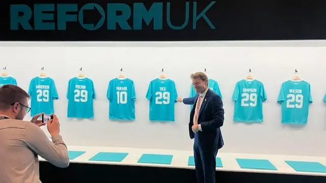Richard Tice stands in front of several Reform UK football shirts. The backs fo the shirts are visible with all of the team bearing number 29, with the exception of the Nigel Farage shirt, which carries the number 10.