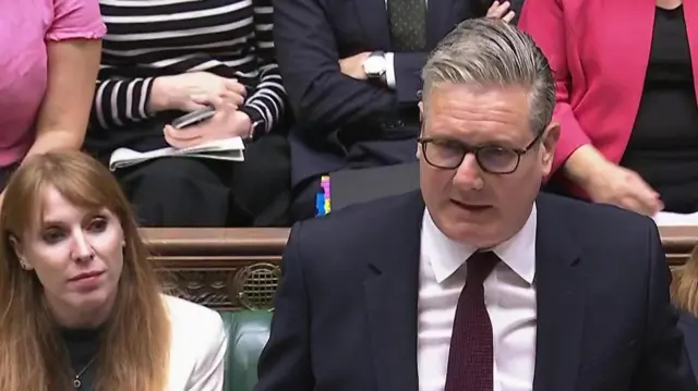 Rayner sits on a bench looking at Starmer who stands in front of her. A row behind them is filled with people but the picture is cropped so you cannot see their heads