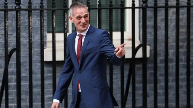 Peter Kyle points to someone off-camera and smiles as he walks past the fence in front of the brick wall of Number 10