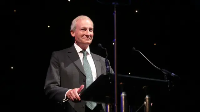 Magnus standing holding a lectern on one side, which has two microphones attached to it at the back of it pointing towards him. The background is black with some twinkling lights. Magnus smiles and looks ahead of him, as he is lit up.