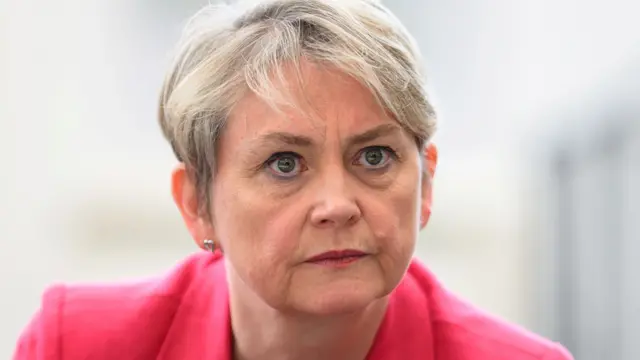 Cooper, who has short cropped blonde hair, wears a bright pink blazer and has a serious expression as she looks on during a visit to a library
