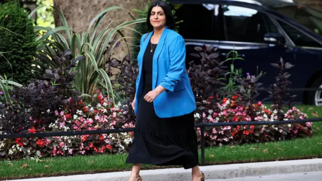 Shabana Mahmood wearing a blue blazer, black dress and leopard print heels, as she smiles at reporters on her way into Downing Street