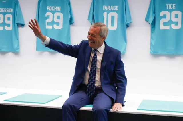 Nigel Farage sits on a bench designed to look like a football team's dressing room. Several of the Reform UK football shirts are hung behind him. Farage is laughing and waving.