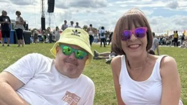 Chris and Claire Jones sit on the grass at the festival. They are wearing colourful sunglasses and white tops. Chris has a yellow bucket hat on.