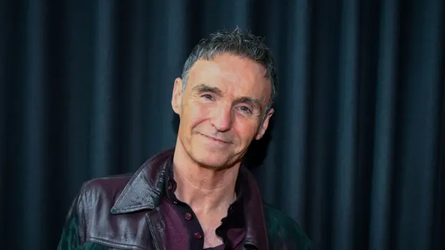 Marti Pellow smiles at the camera in front of navy coloured curtain. He has short dark hair and wears a leather jacket with a dark red top underneath.