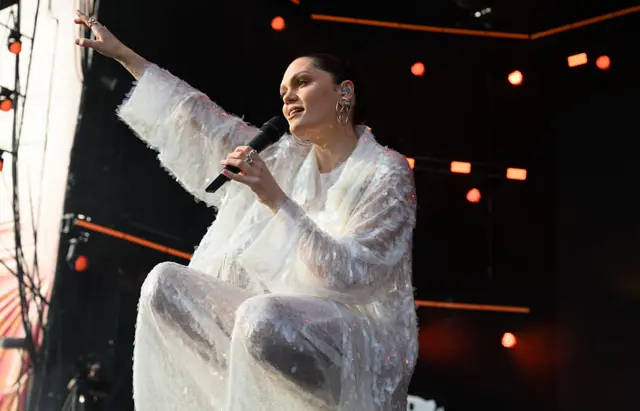 Jessie J knees on stage as she sings into a microphone. One of her arms is raised in the air.