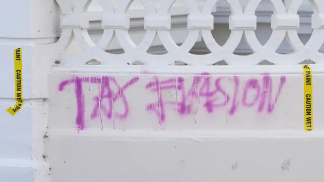 Graffiti reads 'tax evasion' on a white wall near Rayner's property