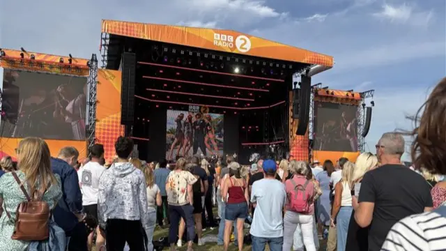 Crowds of people watch a band performing on a large stage in front of them.