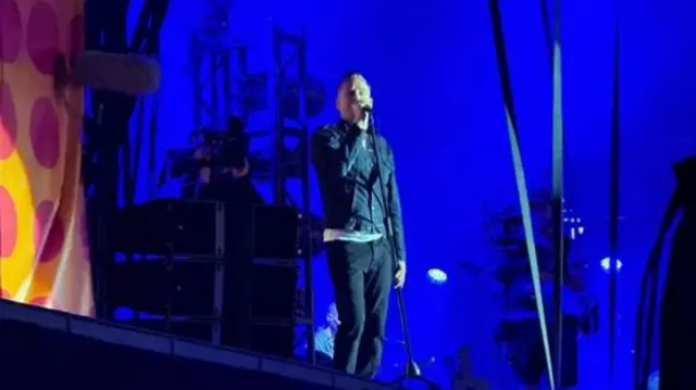 Bryan Adams sings into a microphone on a stage. He wears a jacket and dark trousers.