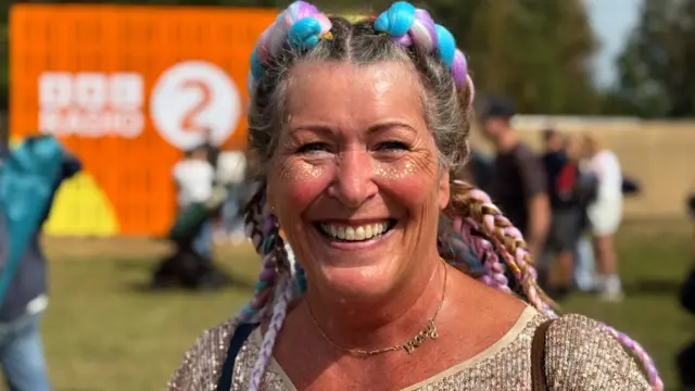 Tracy Crone is smiling and wearing a golden top. She has pink, purple and blue plaits.