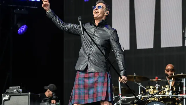 Marti Pellow performs on stage and smiles. He holds a microphone stand in one hand and is raising his other arm. His band plays behind him.