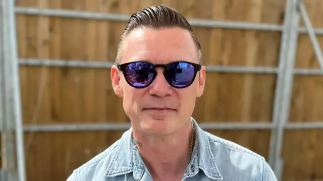 Richard Jones is wearing sunglasses and a denim jacket. He is sitting on a bench in front of a fence and smiling.