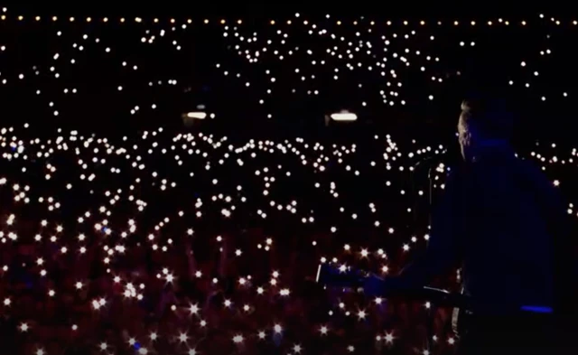 Bryan Adams performs to a crowd a night who have their phone torches on. He is pictured from behind so the crowd can be seen.