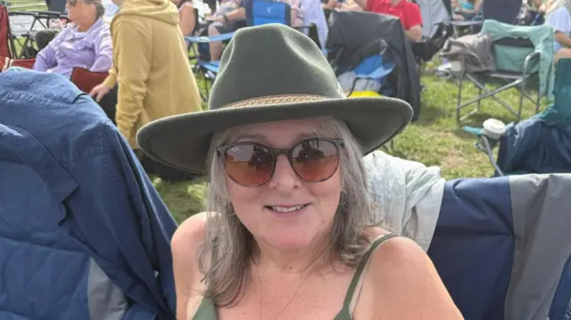A woman sits in a camping chair wearing a green cowboy hat, green cami vest top and sunglasses. She has shoulder length grey hair that falls from beneath her hat.