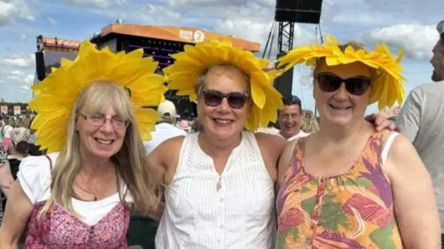 Three women wearing matching sunflower hats smile at the camera during a festival.