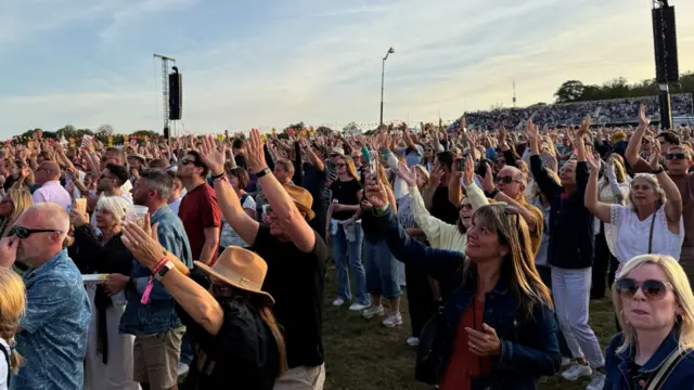 Another view in front of a large crowd of people with their arms raised in the area during a festival.