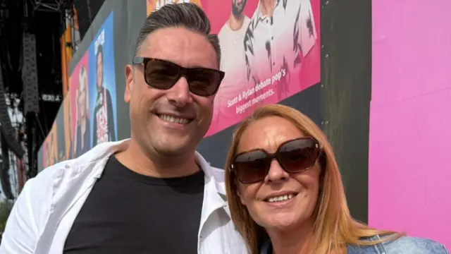 A man with dark hair, sunglasses, a white shirt and black T-shirt underneath, stands next to a woman with red hair, sunglasses and a denim jacket.