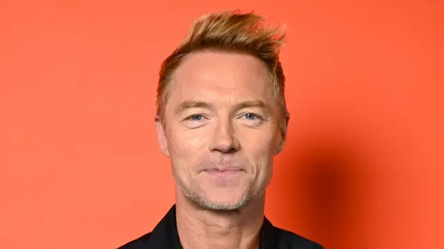 Ronan Keating smiles at the camera in front of an orange backdrop. He has short blonde hair and wears a black shirt.
