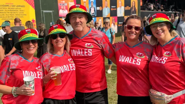 A group of four women and one man in the middle wearing matching Welsh football tops. They are all wearing matching hats.