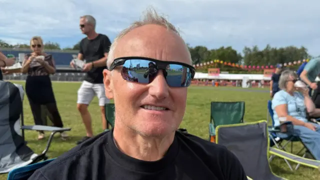 A man with thin grey hair, sunglasses and a black T-shirt smiles at the camera while sitting on a camping chair.