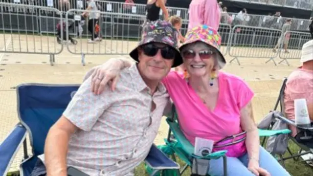 A couple sitting on camping chairs smile at the camera. The woman has her arm around the man next to her. They both wear bucket hats and sunglasses. The man wears a patterned shirt and blue shorts. The woman wears a pink T-shirt and blue trousers.