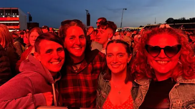 Four women stand together under the glow of an orange stage light and smile at the camera.