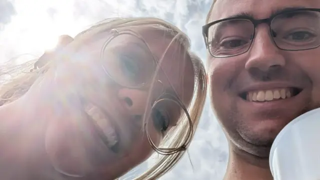 A young woman with blonde hair and glasses and a young man also wearing glasses take a selfie from below. The man holds a pint glass.
