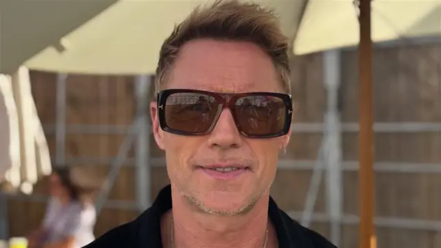 Ronan Keating smiles at the camera backstage. He wears large sunglasses and a black shirt. He has short light coloured hair.