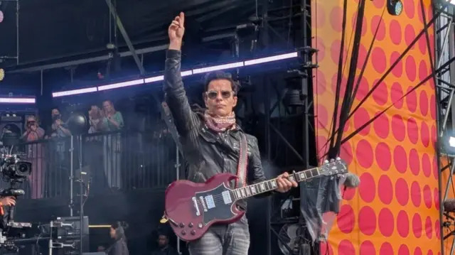 The Stereophonics frontman stands on stage with one hand raised in the area and the other holding his guitar. He wears glasses, jeans and a leather jacket. He has short dark hair.