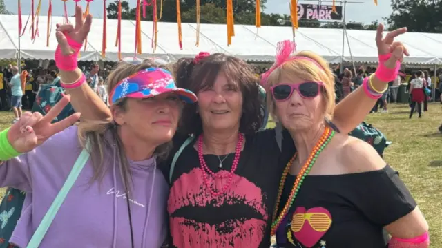 Shirley Nimmo, Janet Barrack and Pauline Haynes all standing arm in arm. They are wearing colourful outfirs including hats and sunglasses. Behind them is a large marquee full of people.