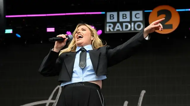 Ella Henderson sings into a microphone and holds one arm out to the side of her.