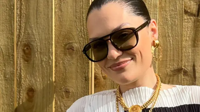 Jessie J stands in front of a wooden fence and smiles at the camera. Her dark hair is tied back and she wears sunglasses, gold chains and a black and white dress.