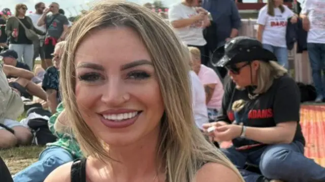 A young woman with long blonde hair smiles at the camera. She has a nose piercing, wears a necklace and wears a black vest top. People sitting on a picnic blanket can be seen behind her as well as other people leaning on a barrier.