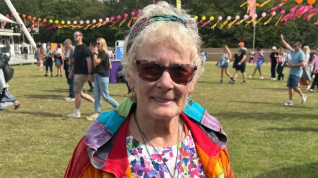 Wanda Fletcher is wearing a multi-coloured jacket over a white, purple and pink T-shirt. She has a bandana in her hair and is wearing sunglasses.