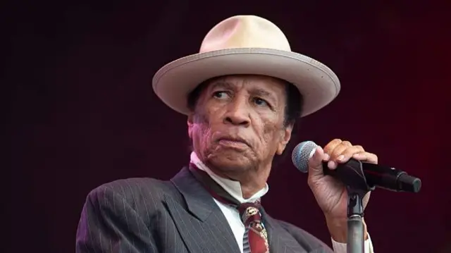 Kid Creole performs on stage and holds a microphone in one hand as he looks to his right. He wears a large hair, suit and tie.