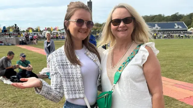 Jennie and Lauren Manley standing in a busy field at Hylands Park. They are both wearing white and sunglasses. They are smiling.