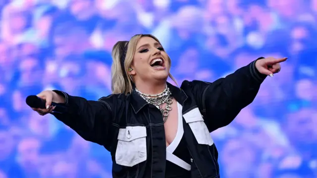 Ella Henderson performs on stage and holds both her arms out from her body. She wears a denim jacket with black top underneath.
