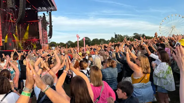 A large crowd of people all with their hands in the air watch Ronan Keating perform on a large stage.