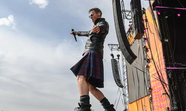 Marti Pellow walks on a stage during a performance. He wears a kilt, boots and leather jacket.