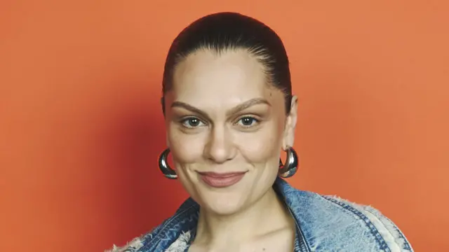 Jessie J stands in front of an orange background and smiles at the camera. Her dark hair is slicked back behind her head. She wears silver hoop earrings and a denim jacket.
